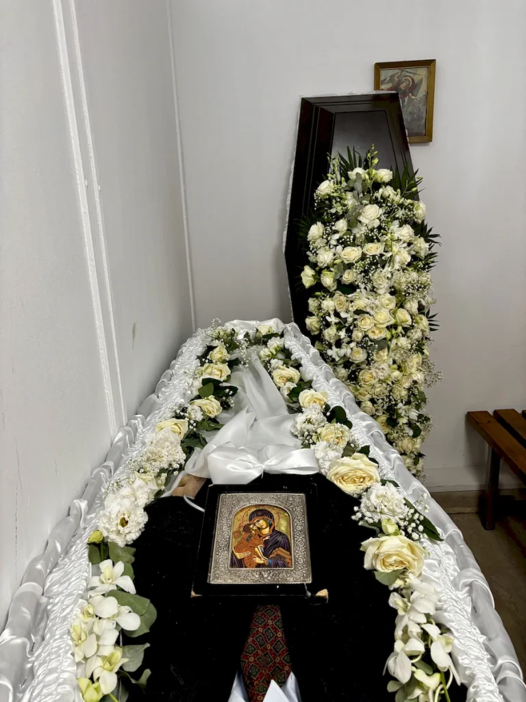 Φωτογραφία με πολυτελή στολισμό καπακιού φέρετρου και μινιμαλιστικό στολισμό σωρού, διακοσμημένα με λευκά άνθη.