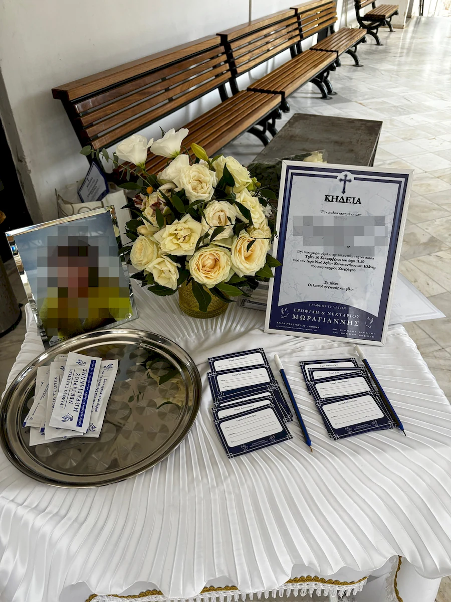 Προσεγμένα διαμορφωμένο τραπεζάκι σε τελετή, με ανθοσύνθεση λευκών λουλουδιών, κορνίζα με φωτογραφία του εκλιπόντος (θολωμένη για ιδιωτικότητα), κηδειόχαρτο, συλλυπητήριες κάρτες με στυλό και μαντιλάκια για τους παρευρισκόμενους.